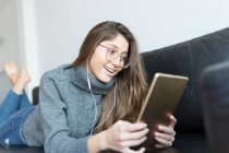 Portrait of smiling young woman lying on couch using earphones and digital tablet — Stock Photo