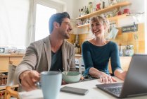 Happy couple sitting at table in kitchen using laptop — Stock Photo