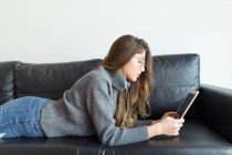 Young woman lying on black leather couch using earphones and digital tablet — Stock Photo