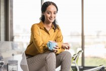 Porträt einer lächelnden jungen Geschäftsfrau, die auf dem Schreibtisch im Büro sitzt und eine Kaffeepause einlegt — Stockfoto