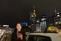 Jovem mulher usando smartphone ao lado de um táxi na cidade à noite, Frankfurt, Alemanha — Fotografia de Stock