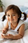 Portrait of smiling little girl with smartphone lying on bed — Stock Photo