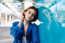Jeune femme au look urbain assise sur le sol et appuyée sur un mur de verre coloré avec son reflet — Photo de stock
