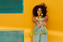 Porträt einer glücklichen jungen Frau in Overalls mit Blumen vor gelber Wand — Stockfoto
