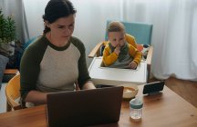 Mujer trabajando en el ordenador portátil en casa mientras su hijo pequeño viendo películas en el teléfono móvil - foto de stock