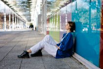 Jeune femme au look urbain assise sur le sol et appuyée sur un mur de verre coloré avec son reflet — Photo de stock