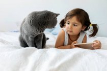 Portrait of little girl lying on bed with smartphone looking at grey British shorthair — Stock Photo