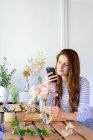 Spain, Portrait of young beautiful redhead woman using smart phone while potting plants at table — Stock Photo