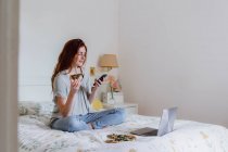 Belle femme utilisant un téléphone mobile tout en étant assis avec pizza et ordinateur portable sur le lit à la maison — Photo de stock