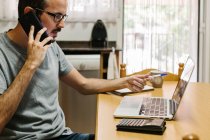 Homem adulto médio usando telefone inteligente enquanto trabalhava no laptop em casa — Fotografia de Stock