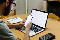 Homem adulto médio usando telefone inteligente e laptop enquanto trabalhava em casa — Fotografia de Stock