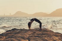 Femme mûre pratiquant le yoga à la plage rocheuse le soir, pose de l'arc — Photo de stock