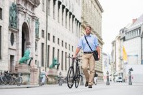 Smiling businessman carrying messenger bag walking with bicycle on street in city — Stock Photo
