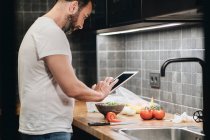 Reifer Mann, der in der Küche steht, Essen zubereitet, Online-Rezept verwendet — Stockfoto