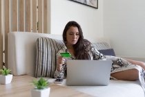Beautiful young woman holding credit card for online shopping through laptop while lying on sofa at home — Stock Photo
