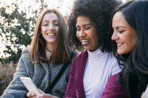 Cheerful young woman showing smart phone to female friends while sitting in park — Stock Photo
