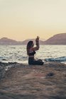 Femme mûre pratiquant le yoga à la plage de rochers le soir — Photo de stock