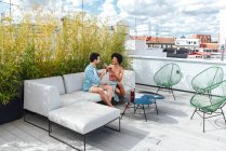 Multi-ethnic couple toasting red wine glasses while sitting on sofa at penthouse terrace — Stock Photo