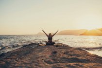 Femme mûre pratiquant le yoga à la plage de rochers le soir — Photo de stock