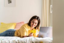 Smiling woman looking at credit card while using laptop in bedroom — Stock Photo