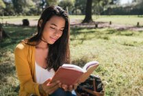 Giovane donna lettura libro mentre seduto su terreni erbosi nel parco — Foto stock