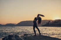 Femme mûre pratiquant le yoga à la plage rocheuse pendant le coucher du soleil — Photo de stock