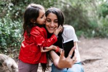 Figlia baciare la madre mentre prende un selfie nel bosco — Foto stock