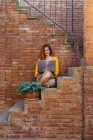 Jeune femme utilisant un ordinateur portable tout en étant assis sur un escalier contre un mur de briques — Photo de stock