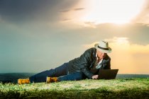 Homem sênior usando laptop enquanto estava deitado em campo durante o pôr do sol — Fotografia de Stock