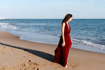 Delicate woman in red dress walking at the sea, feeling the sun — Photo de stock