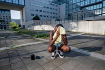 Young woman wiping head while crouching by smart phone and speaker on footpath in city — Photo de stock