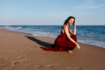 Delicate woman in red dress sittingh on the beach, feeling the sun — Photo de stock