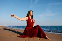 Delicate woman in red dress sittingh on the beach, feeling the sun — Photo de stock