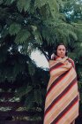 Donna con gli occhi chiusi avvolto in coperta ascoltare musica attraverso le cuffie da albero acacia dealbata — Foto stock