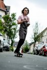 Smiling male barista holding coffee cup while skateboarding on road in city — Stock Photo