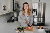 Femme souriante hachant des légumes sur la planche à découper dans la cuisine — Photo de stock
