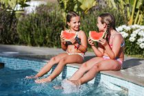 Happy twin girls sitting with watermelon slice at poolside on sunny day — Stock Photo