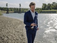 Empresario usando teléfono móvil mientras está de pie en la orilla del río durante el día soleado - foto de stock