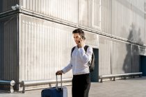 Бизнесмен с чемоданом разговаривает по смартфону, стоя на тропинке напротив здания — стоковое фото