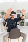 Young man with arms raised using visual reality simulator while sitting on chair at office — Stock Photo