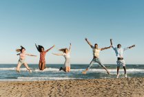 Allegro giovani amici che saltano con la mano alzata sulla spiaggia durante la giornata di sole — Foto stock