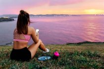 Sportswoman with smart phone sitting on grass while looking at sea against sky during sunset — Stock Photo