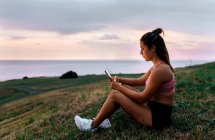 Young female athlete using smart phone while sitting on grass during sunset — Stock Photo