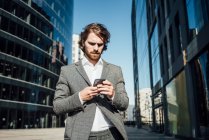 Male professional using mobile phone while standing against modern office building in downtown on sunny day - foto de stock