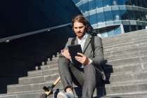 Молодой предприниматель использует цифровой планшет, сидя с электрическим самокатом на ступенях в финансовом районе — стоковое фото
