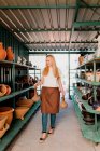 Smiling female owner holding potteries while walking amidst shelves in workshop — Stock Photo