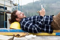 Trabajador de la construcción usando un teléfono inteligente mientras come comida en madera en el sitio de construcción - foto de stock