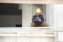 Строитель с помощью цифрового планшета, сидя на ступеньках на стройке — стоковое фото