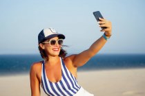 Улыбающаяся женщина делает селфи, сидя на пляже на фоне ясного голубого неба в солнечный день — стоковое фото