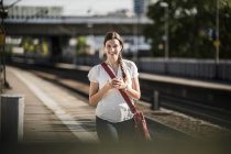 Красивая молодая женщина слушает музыку через мобильный телефон во время прогулки по железнодорожной станции платформы — стоковое фото